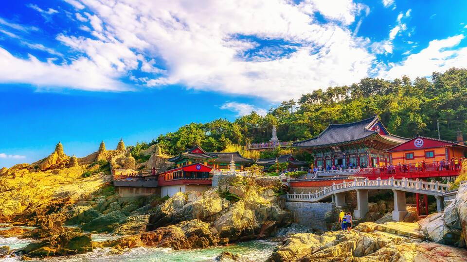 A traditional Chinese temple complex with pagodas, a red-roofed building, and a bridge over a rocky river.