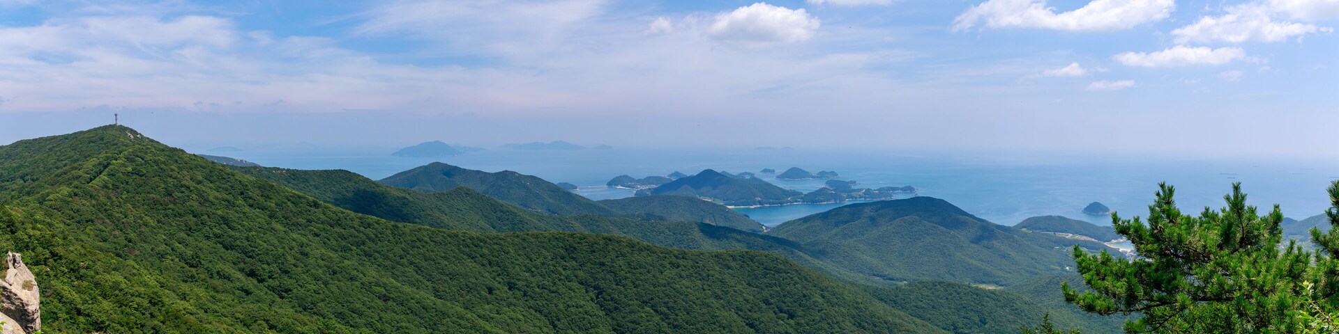 A mountainous landscape with dense green forests, a clear blue sky, and a distant view of a coastline with small islands.
