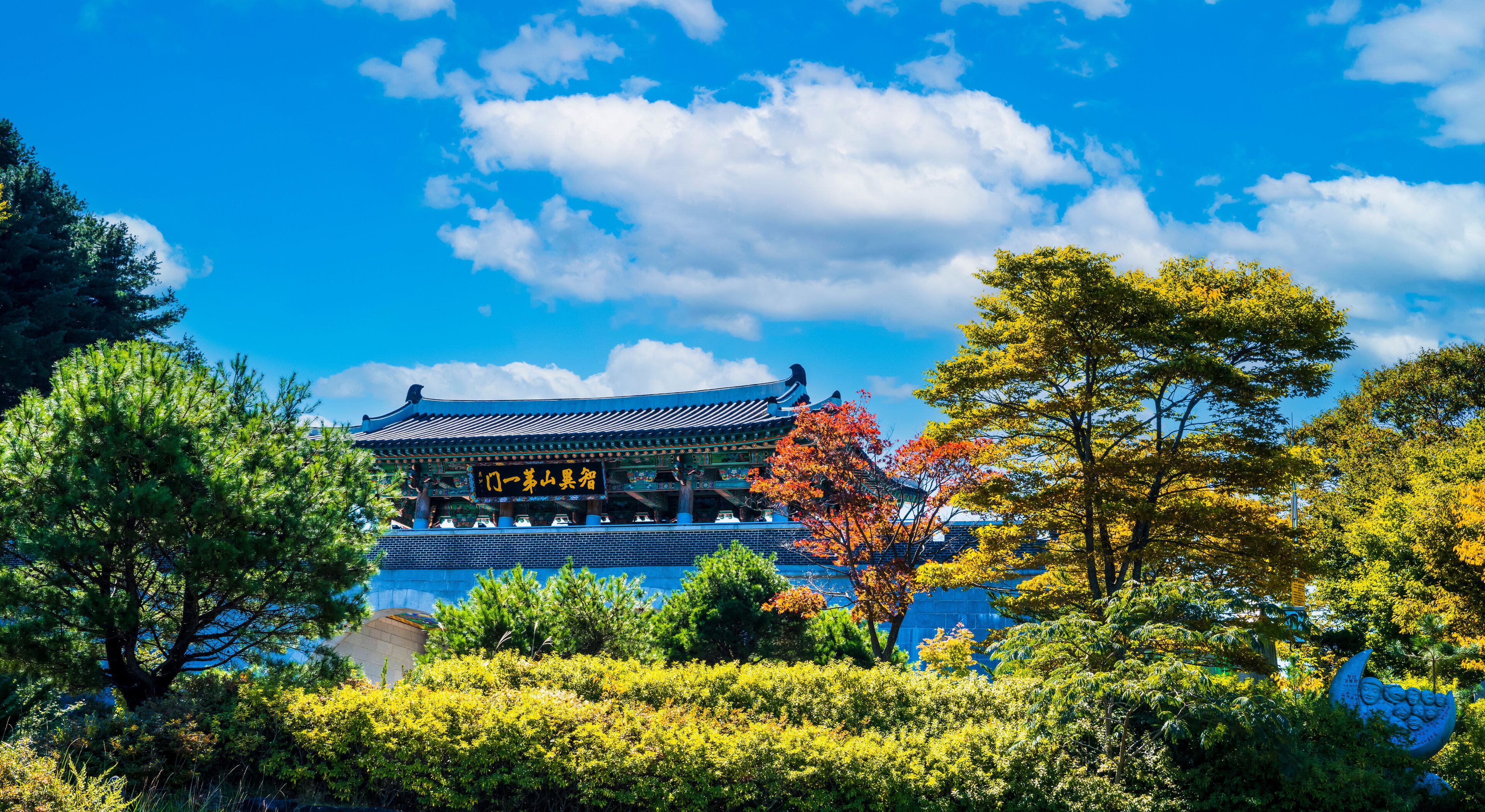A traditional Korean building with a blue roof and golden decorations, surrounded by lush greenery and autumn-colored trees.