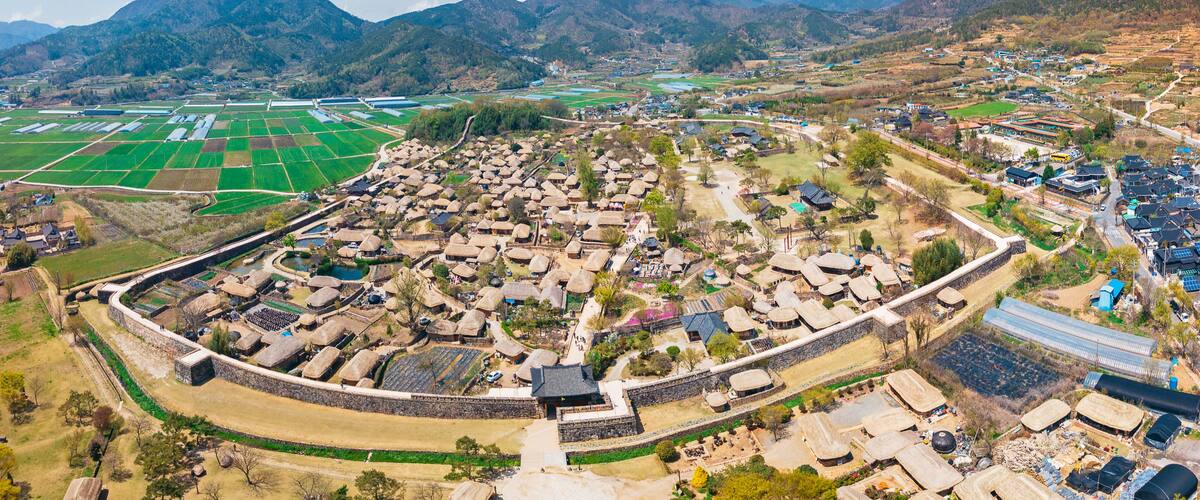 An aerial view of a village with traditional houses, surrounded by fields and mountains.