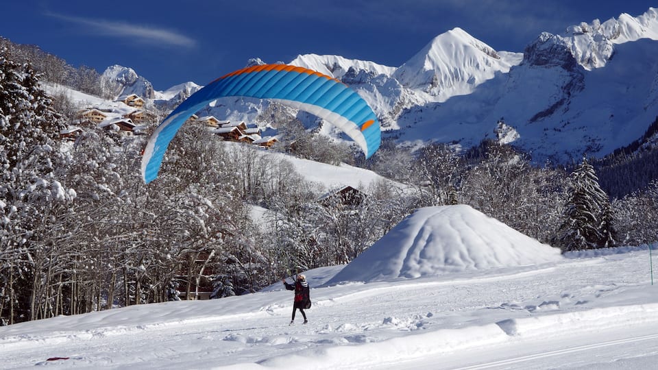 A person paragliding over a snowy mountain landscape with snow-covered trees and houses.
