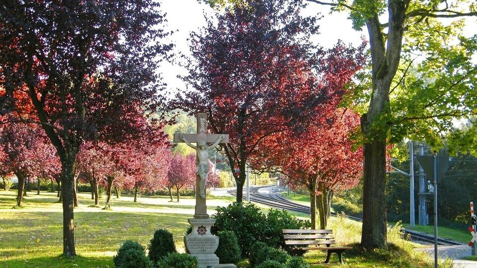 A park with a cross monument, trees with autumn leaves, a bench, and a railway crossing.