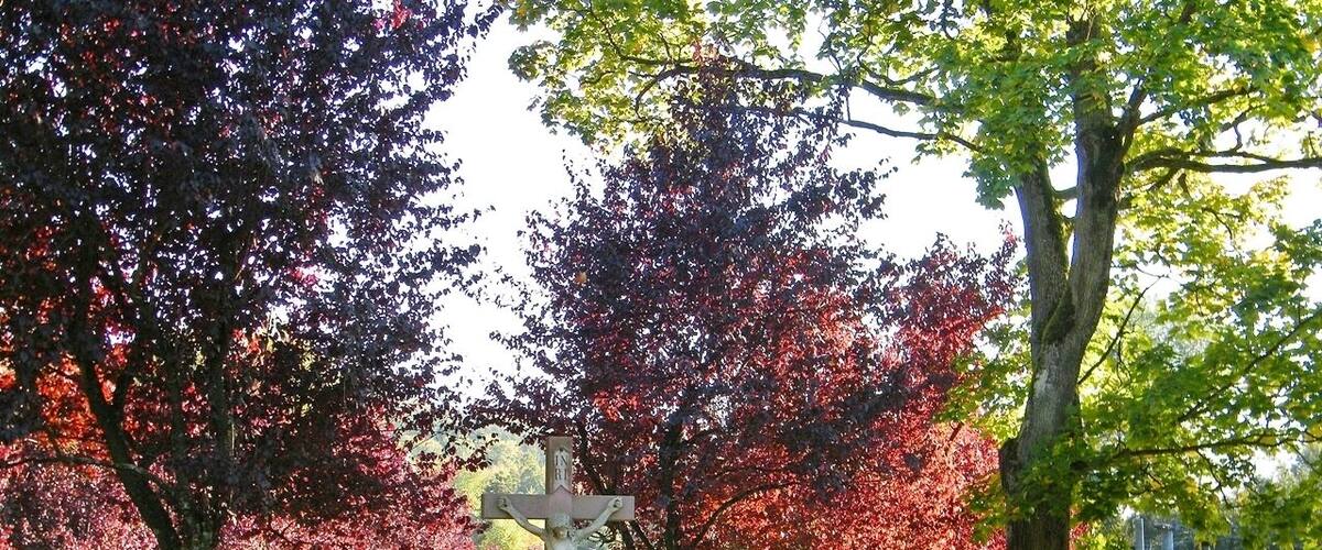 A park with a cross monument, trees with autumn leaves, a bench, and a railway crossing.