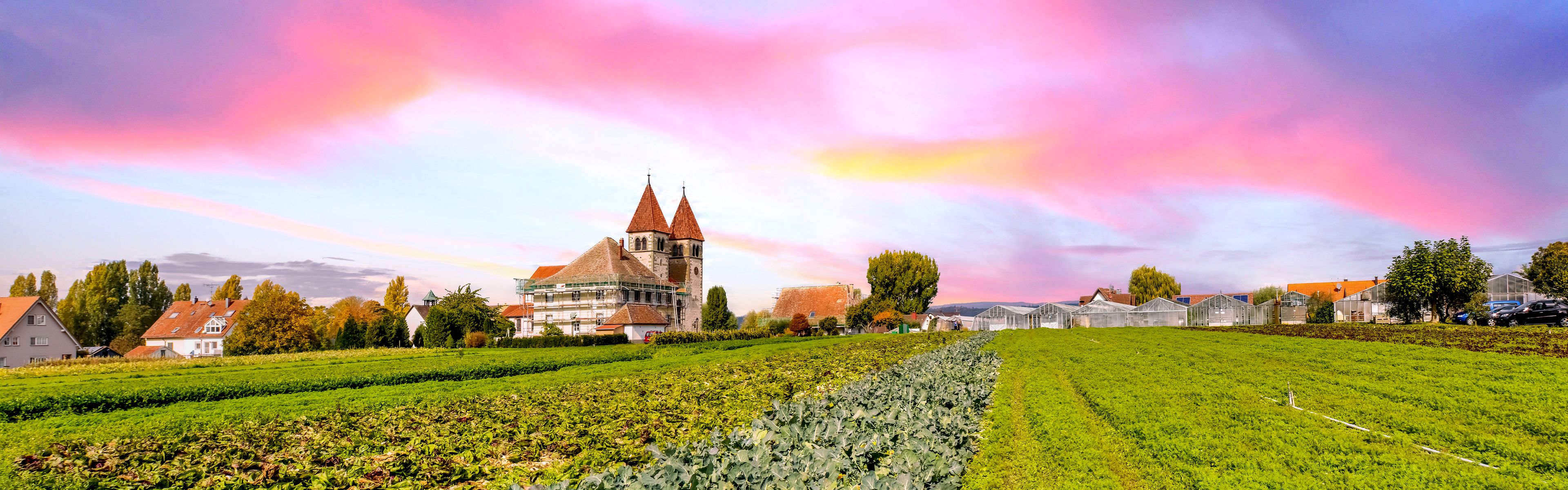 A church with a red roof and a tower, surrounded by green fields and houses.
