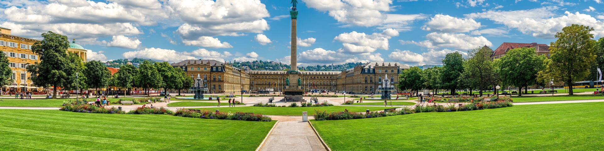 A large green park with a central monument, a fountain, and classical architecture in the background.