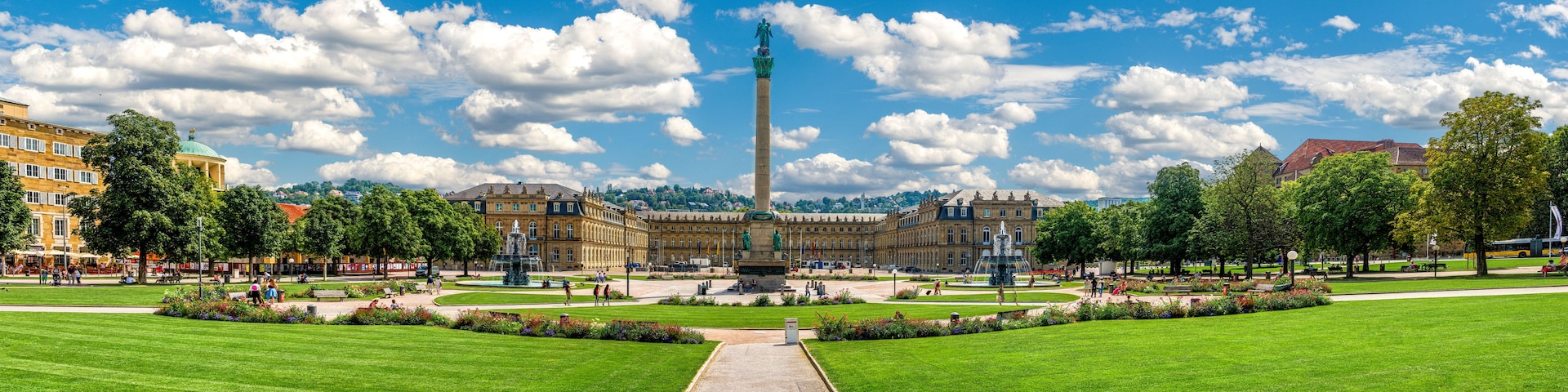 Un gran parque verde con un monumento central, una fuente y arquitectura clásica al fondo.