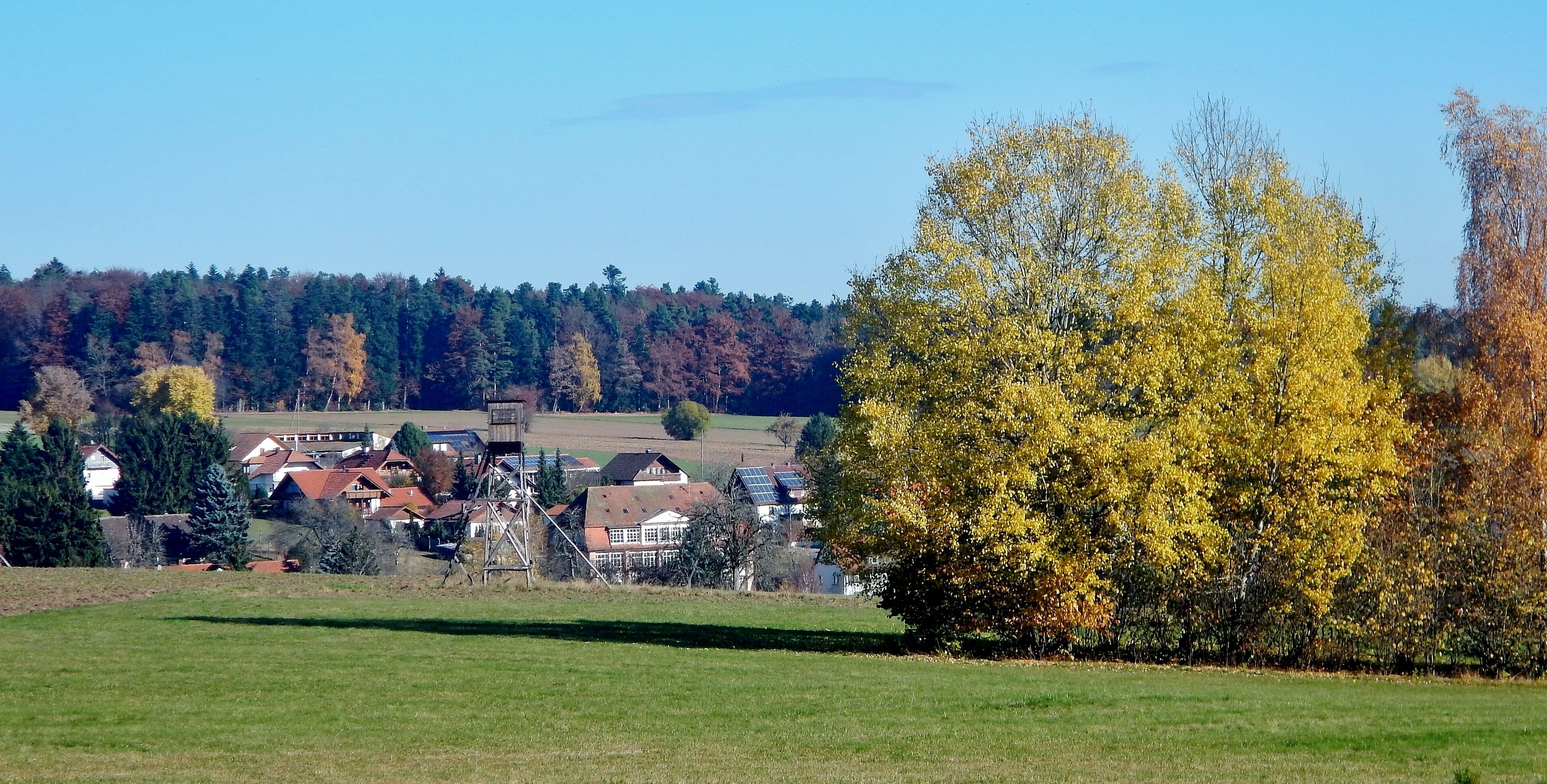 A rural landscape with autumn-colored trees, a village with houses, and a clear blue sky.