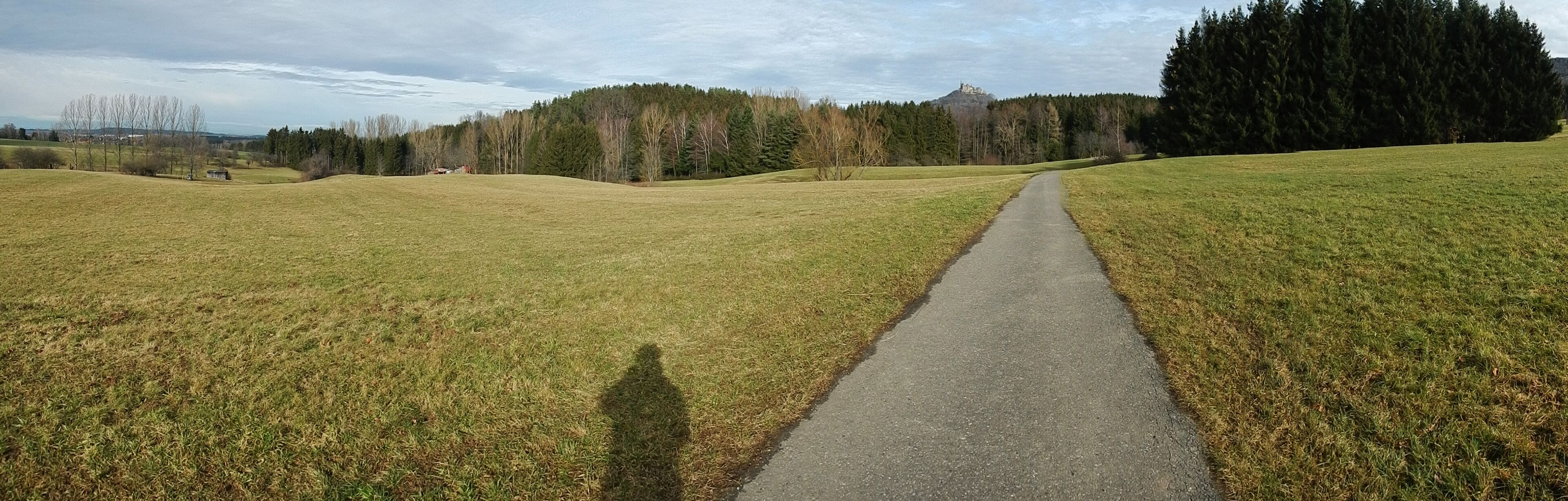 A paved path through a grassy field with a forested hill in the background.