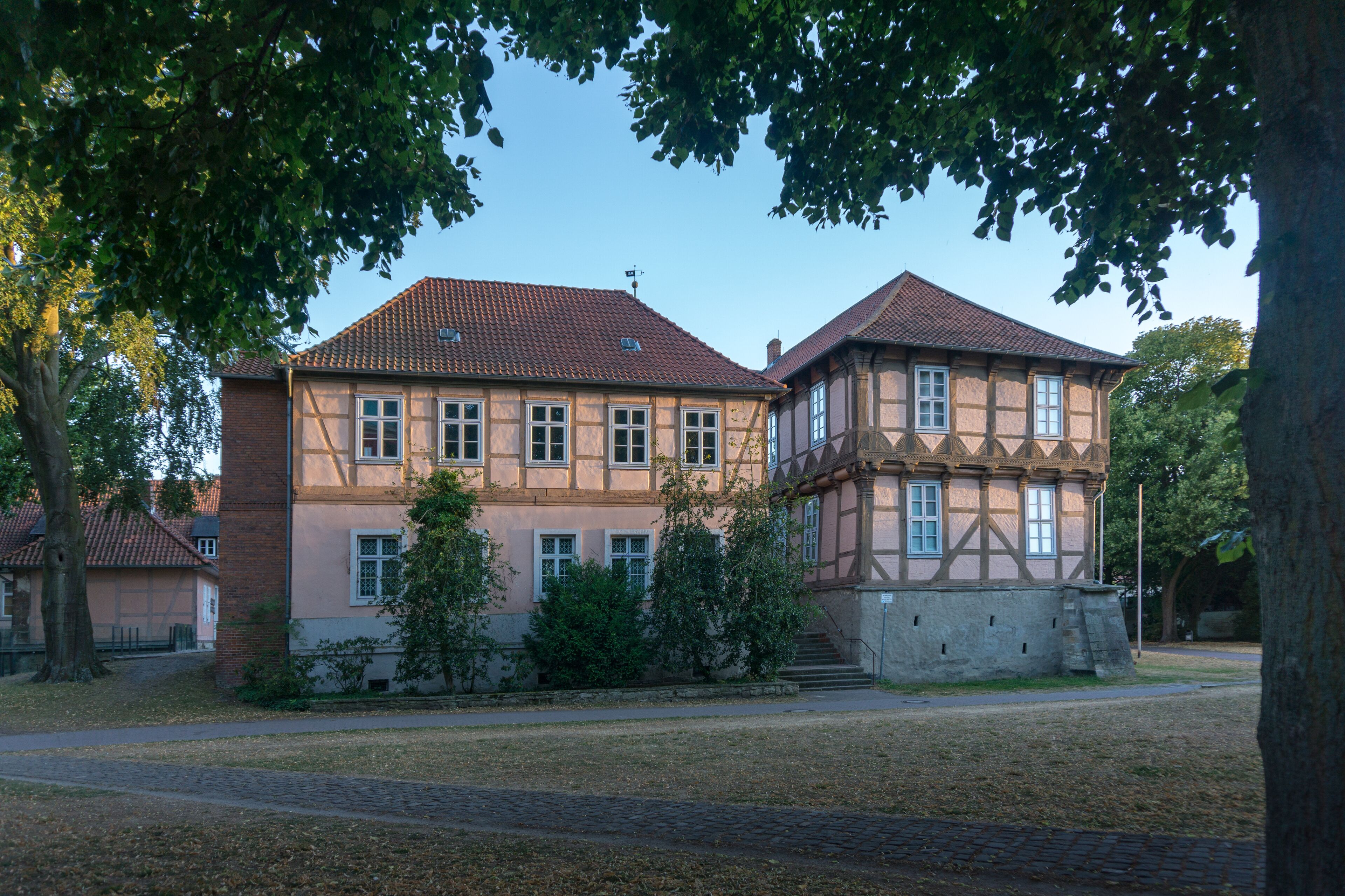 A historic building with a tiled roof and timber framing, surrounded by trees and a cobblestone path.