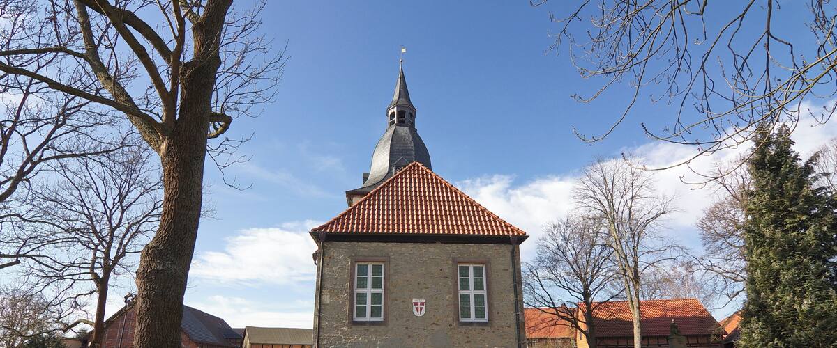A small church with a red-tiled roof and a spire, surrounded by trees and a grassy area.