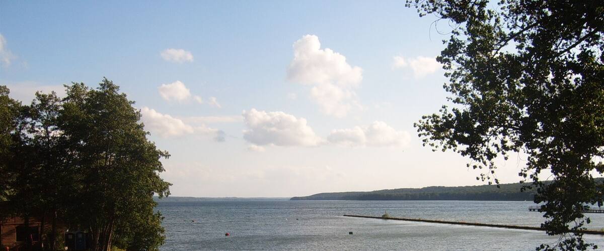 A calm lake with a wooden dock and trees on the left, a distant shoreline with hills, and a clear sky with clouds.
