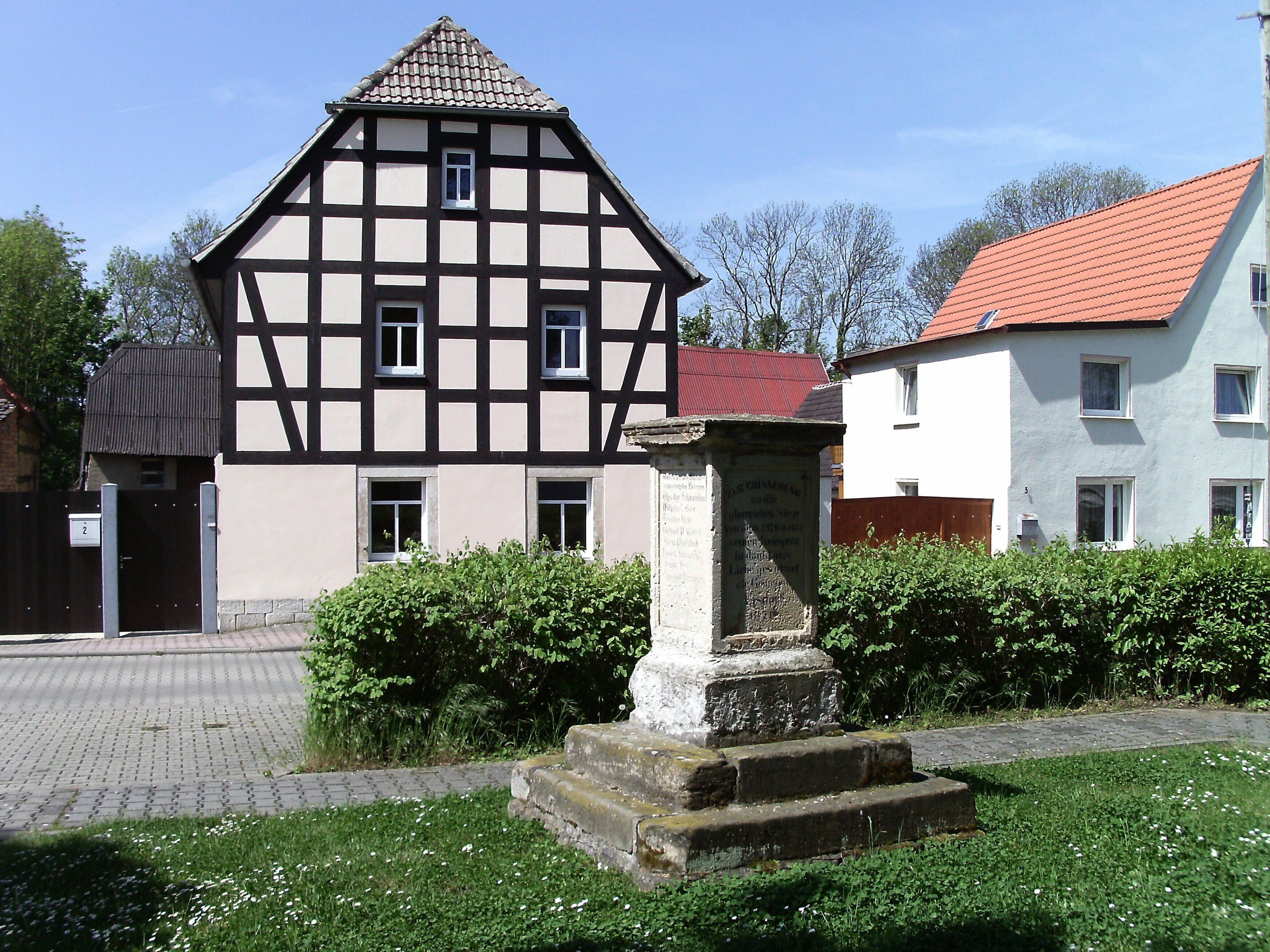 A traditional half-timbered house with a red-tiled roof, a stone monument, and a paved pathway.