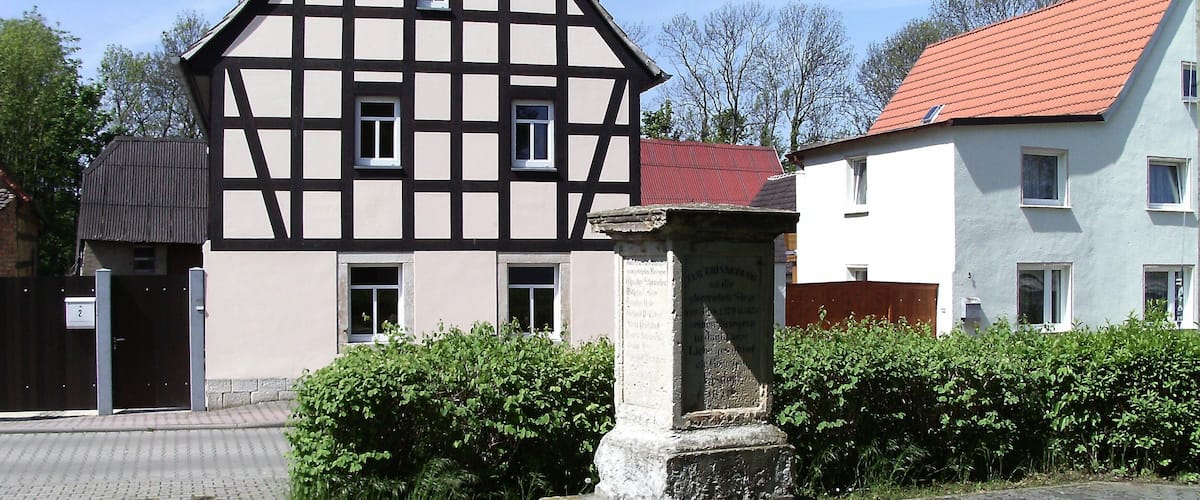 A traditional half-timbered house with a red-tiled roof, a stone monument, and a paved pathway.