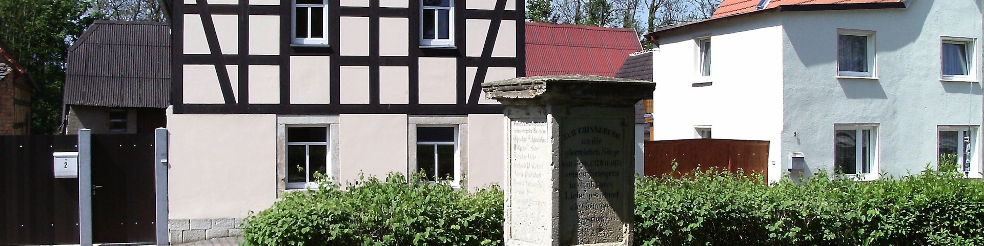 A traditional half-timbered house with a red-tiled roof, a stone monument, and a paved pathway.