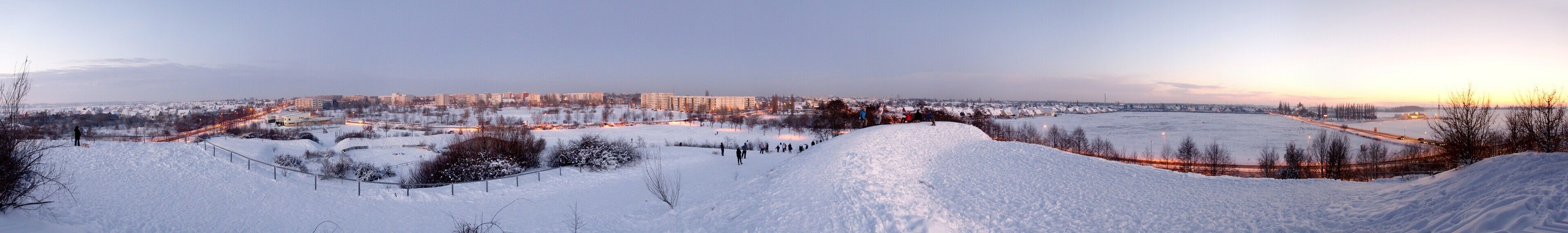 A snowy landscape with a city in the background, a snow-covered hill, and a flat area with trees.