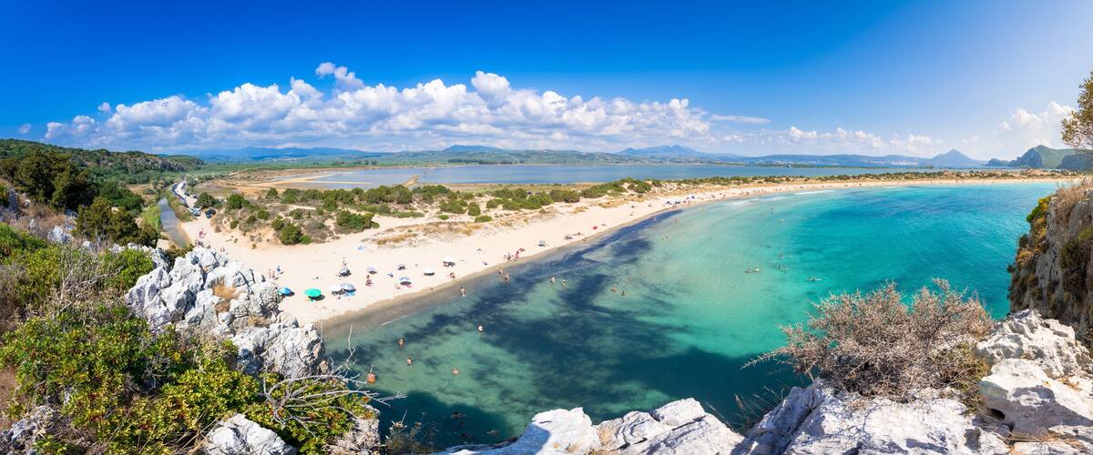 A coastal landscape with a sandy beach, clear turquoise waters, rocky cliffs, and distant mountains under a blue sky.
