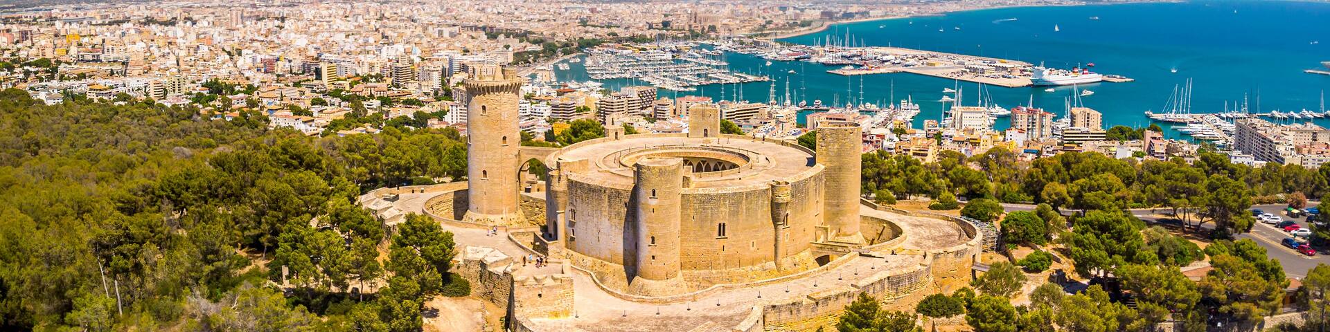 A coastal cityscape with a historic fortress, a marina with numerous boats, and a clear blue sea.