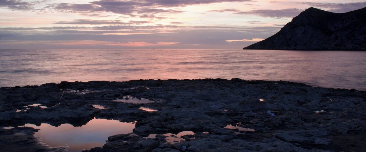 A rocky shoreline with a calm sea and a mountain in the distance during sunset.