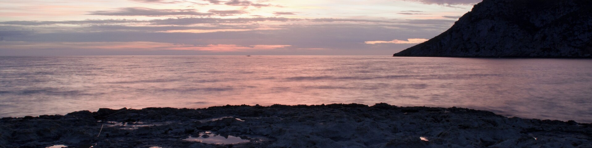 A rocky shoreline with a calm sea and a mountain in the distance during sunset.