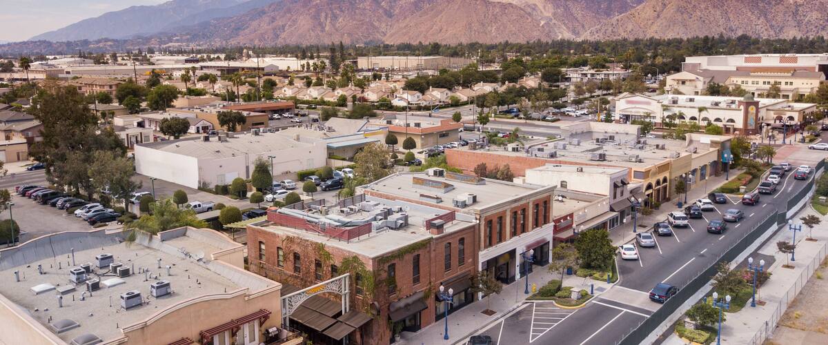 A town with a mountain range in the background, a main street with parked cars, and several buildings with flat roofs.
