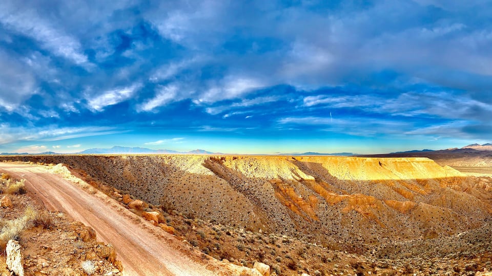 A desert landscape with a winding road, a car, and distant mountains under a partly cloudy sky.