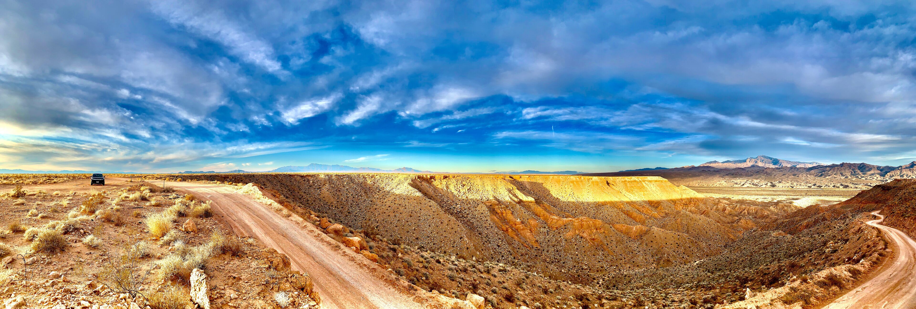 A desert landscape with a winding road, a car, and distant mountains under a partly cloudy sky.