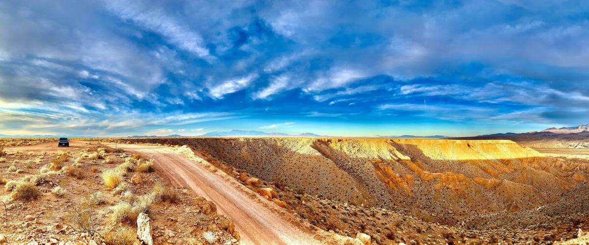 A desert landscape with a winding road, a car, and distant mountains under a partly cloudy sky.