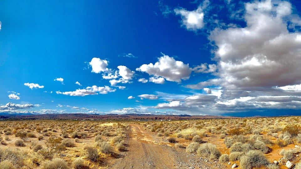 A desert landscape with sparse vegetation, a dirt road, and a yellow house in the distance under a partly cloudy sky.