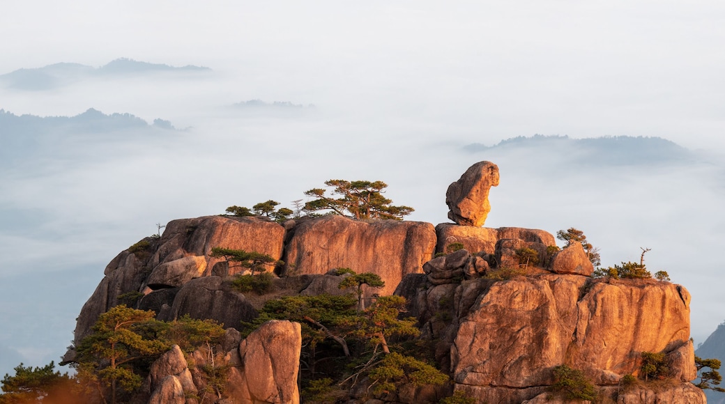 A rocky mountain peak with a prominent rock formation balanced on top, surrounded by trees and shrouded in mist.