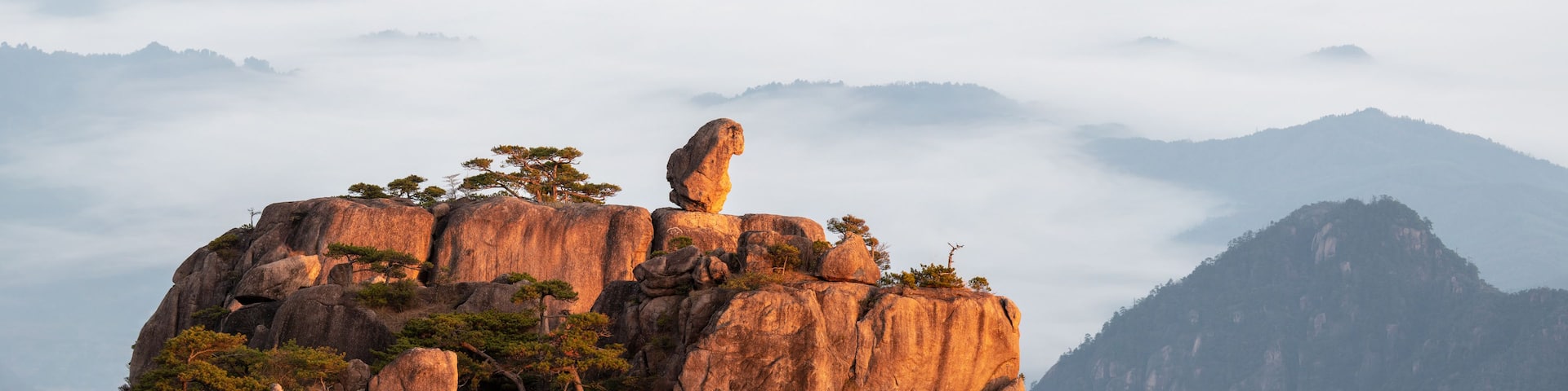A rocky mountain peak with a prominent rock formation balanced on top, surrounded by trees and shrouded in mist.