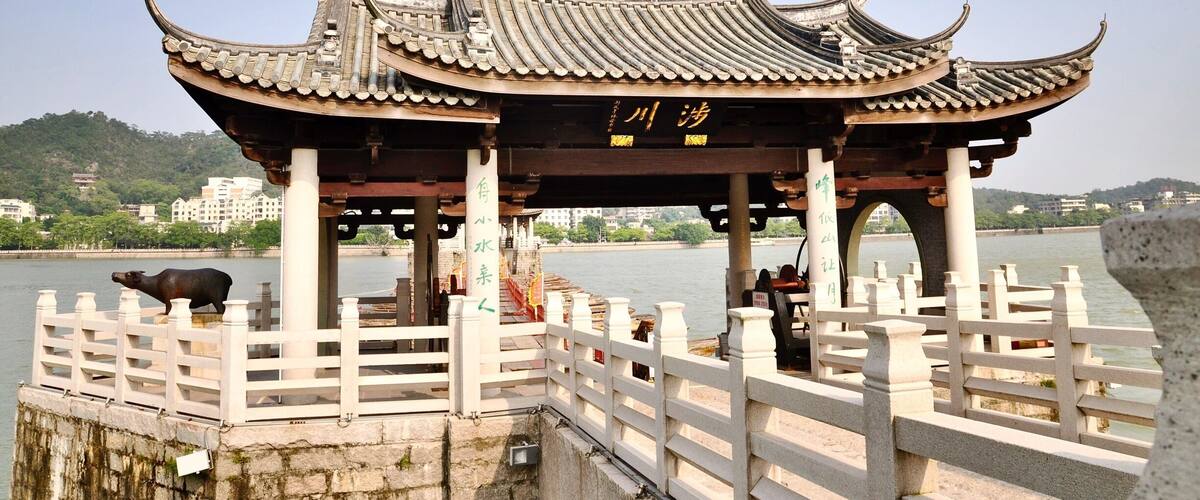A traditional Chinese pavilion by a river with a white fence and a stone statue of a buffalo.