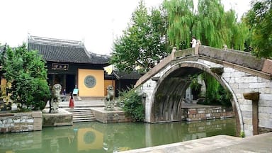 Un jardín chino tradicional con un puente de piedra sobre un estanque, rodeado de árboles y un edificio con tejado de tejas.