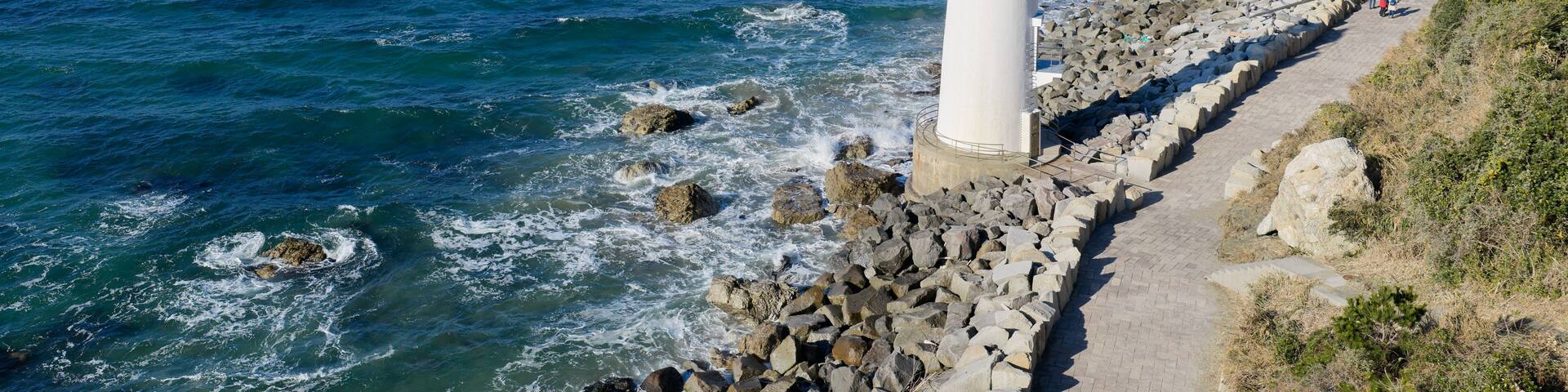 A white lighthouse by the sea, rocky shore, and a clear blue sky.
