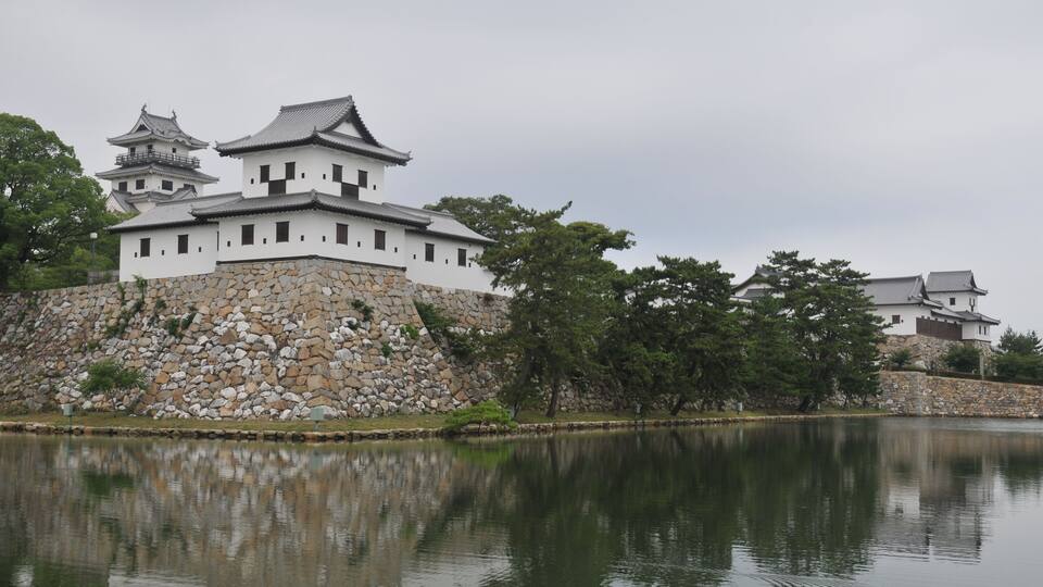 A traditional Japanese castle with multiple white buildings and a stone wall, surrounded by trees and a calm lake.