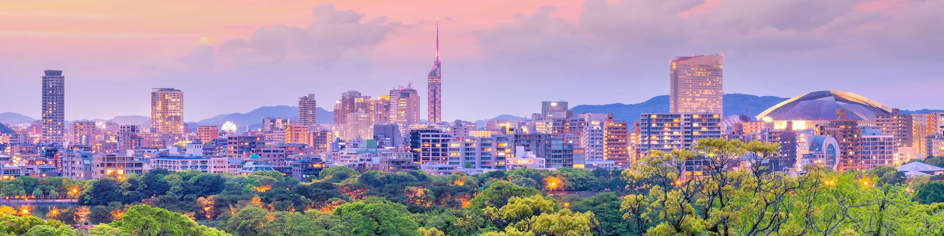 Paisagem urbana com edifícios modernos e uma torre imponente, tudo envolto em vegetação exuberante.