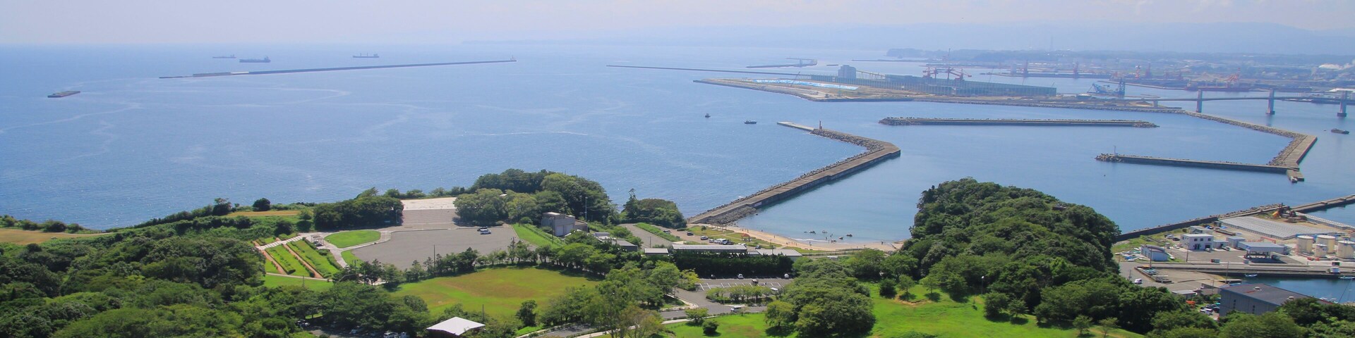 Paisagem costeira com um amplo parque verde, um rotatório e um porto com barcos.