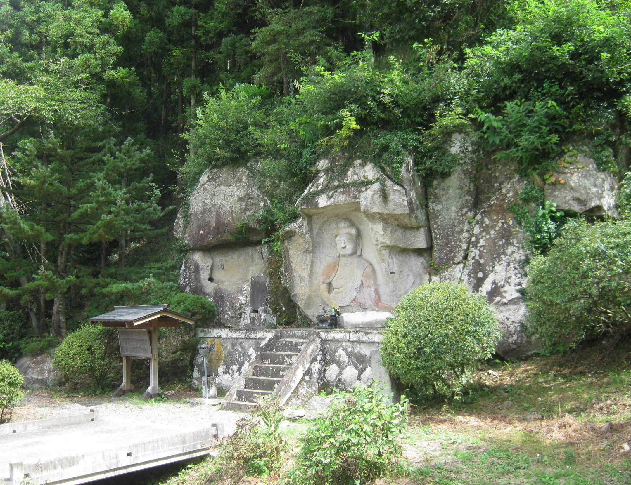 A large Buddha statue carved into a rock, surrounded by lush greenery and a small shrine with steps leading up to it.