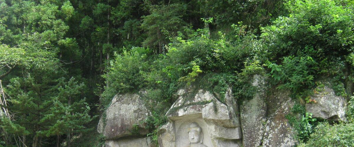 A large Buddha statue carved into a rock, surrounded by lush greenery and a small shrine with steps leading up to it.