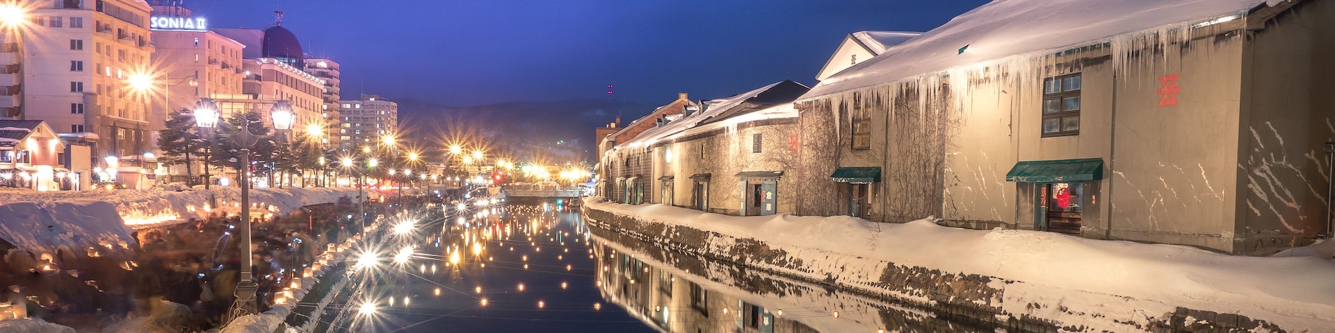 Um canal coberto de neve refletindo luzes da cidade, edifícios com letreiros iluminados e um céu noturno limpo.