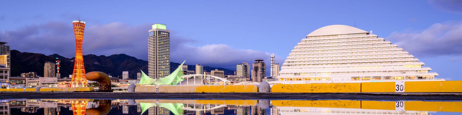 A cityscape with a prominent tower, a modern building, and a large dome structure reflecting on a calm body of water.