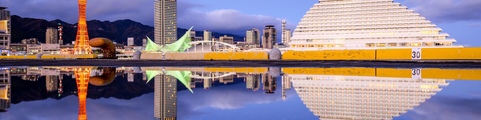 Un paisaje urbano con una torre destacada, un edificio moderno y una gran estructura en forma de cúpula reflejada en una masa de agua tranquila.