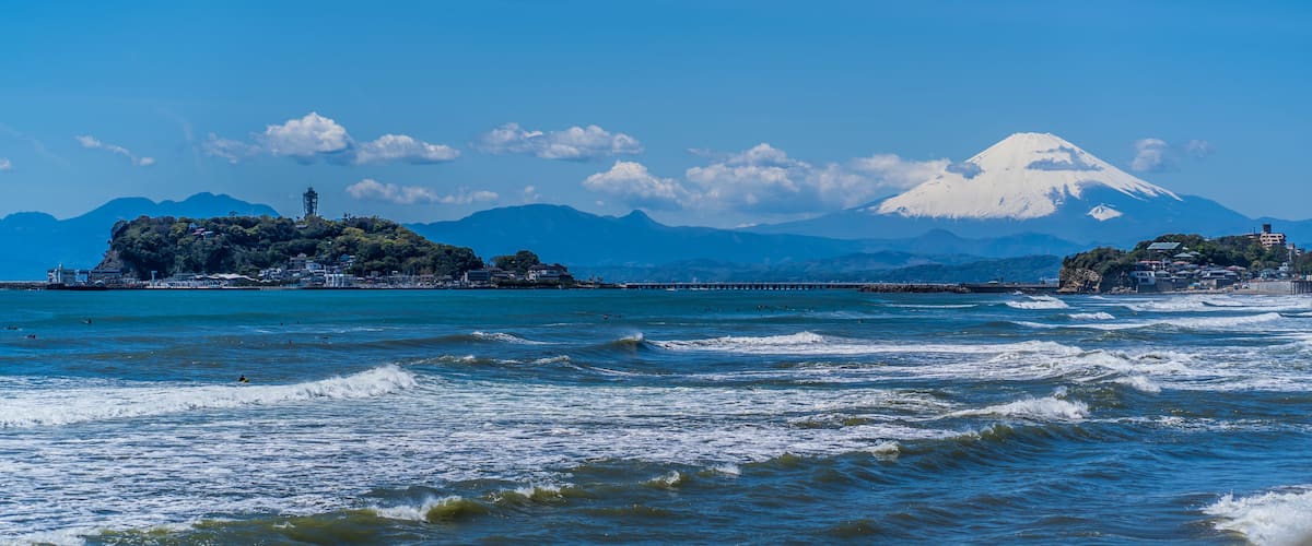 A coastal scene with waves, a distant snow-capped mountain, and a small island with buildings.