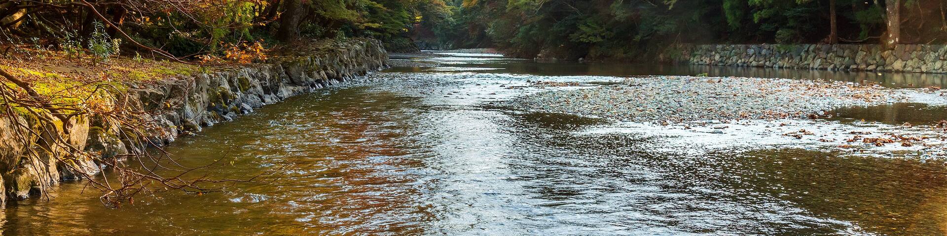 A river flanked by trees with autumn foliage, a stone wall, and a clear sky.