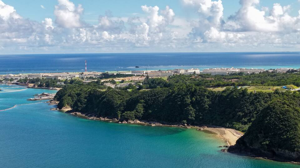 A coastal landscape with a lighthouse, a large building complex, and a forested hill.
