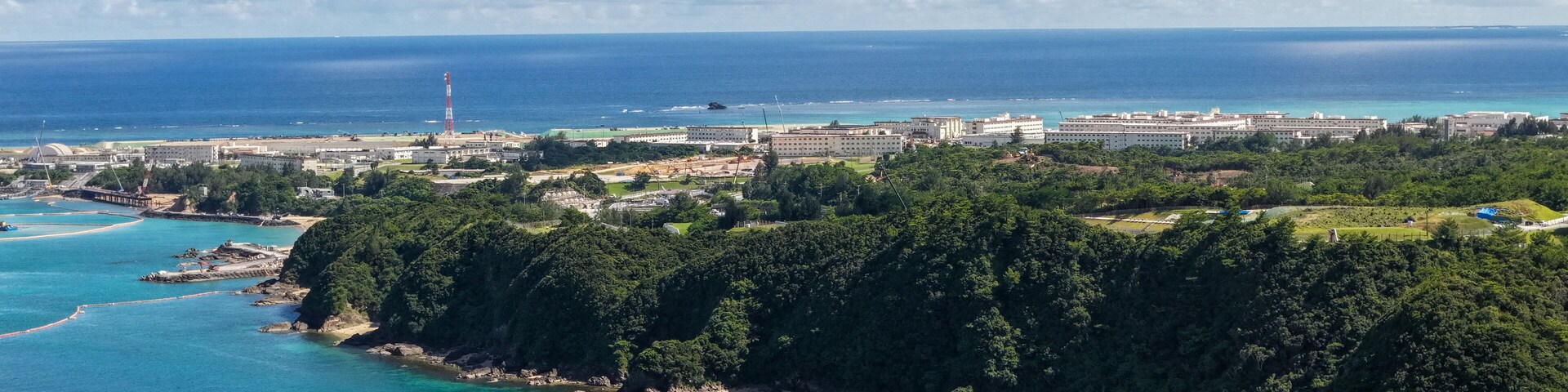 Un paisaje costero con un faro, un gran complejo de edificios y una colina boscosa.