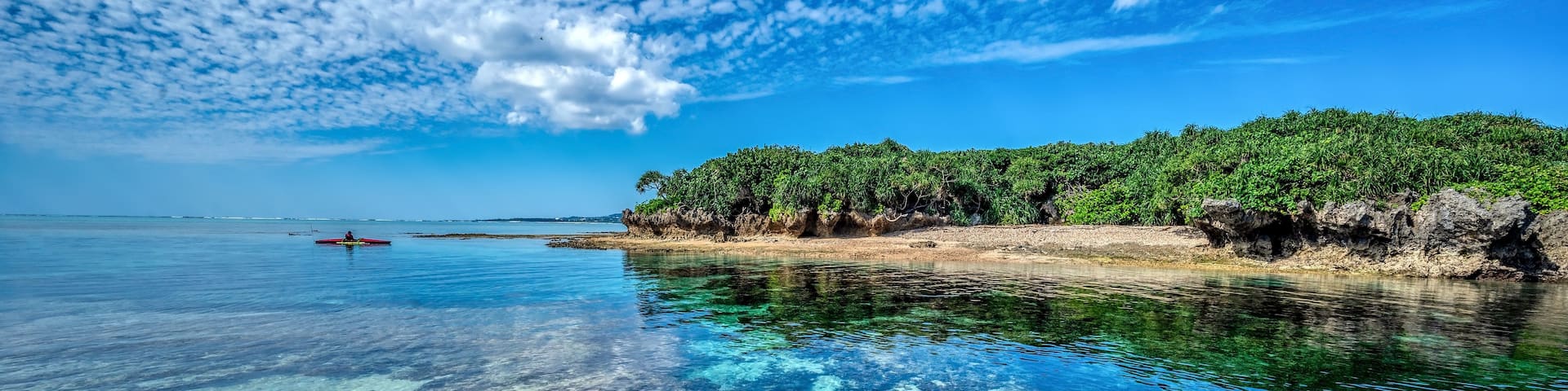 A clear blue sea with a rocky shoreline, a small boat, and a distant island with dense vegetation.