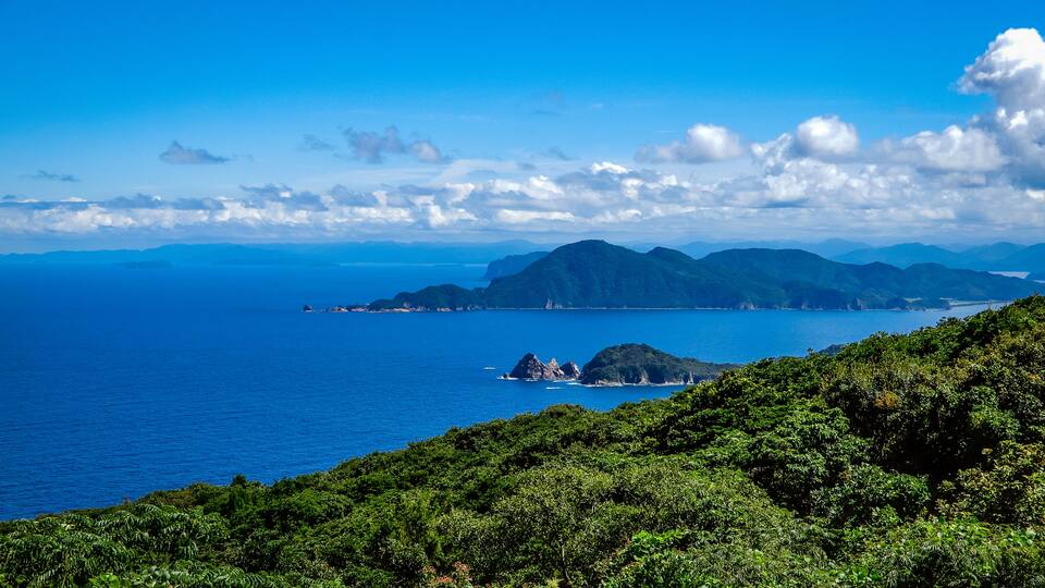 A coastal landscape with a clear blue sea, green hills, and distant mountains under a partly cloudy sky.