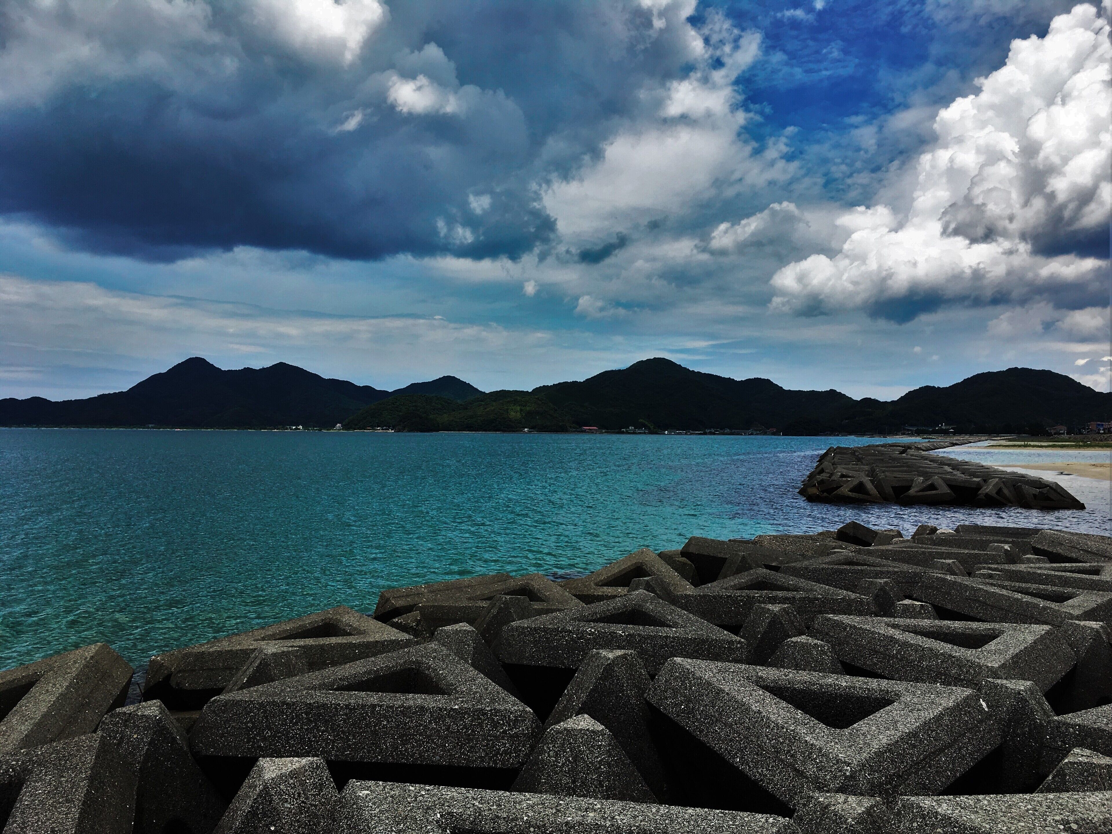 A coastal landscape with a clear blue sea, rocky breakwaters, and a mountainous backdrop under a partly cloudy sky.
