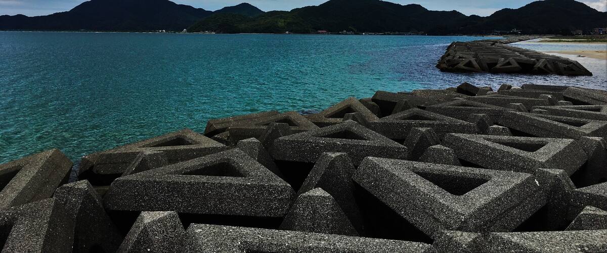A coastal landscape with a clear blue sea, rocky breakwaters, and a mountainous backdrop under a partly cloudy sky.