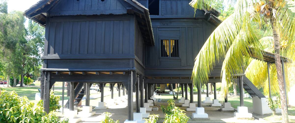 A traditional wooden house with a steep gable roof, supported by concrete stilts, surrounded by palm trees and a well-maintained garden.